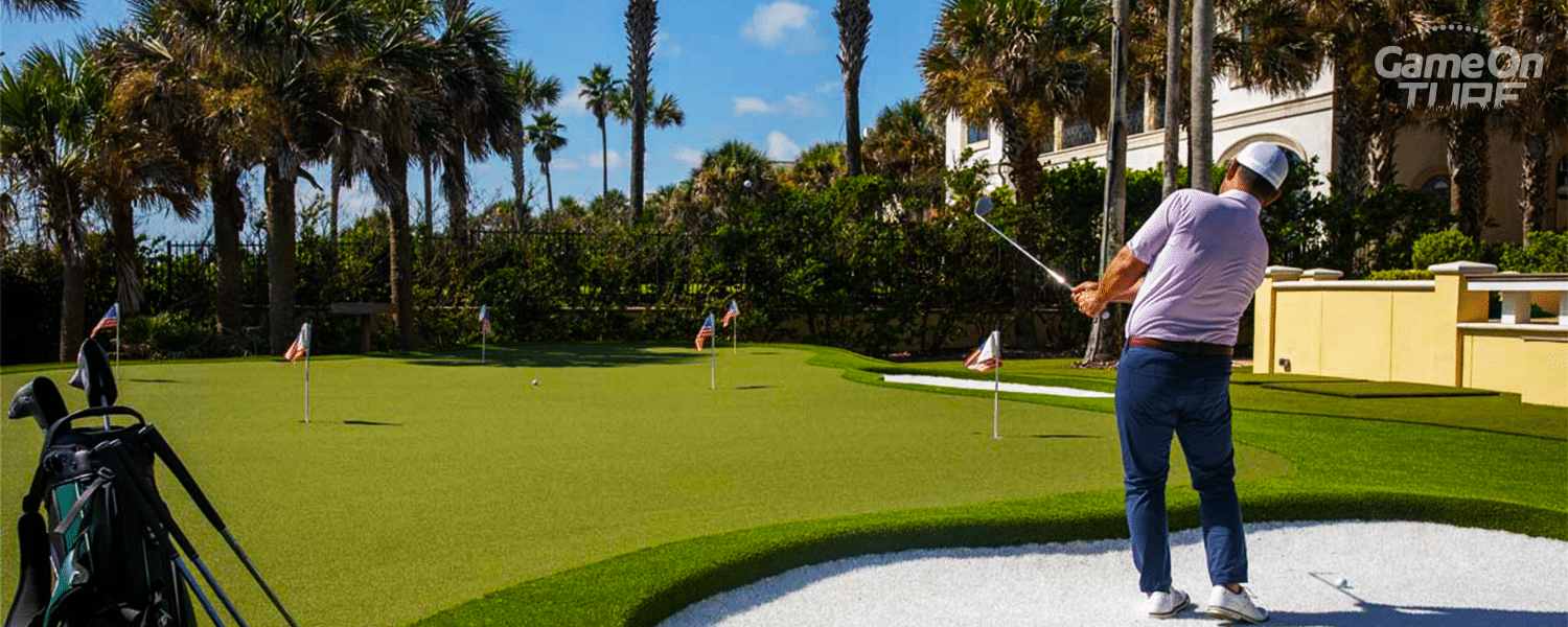 Golfer chipping from a bunker onto an artificial turf putting green in NZ, with flags and palm trees in the background