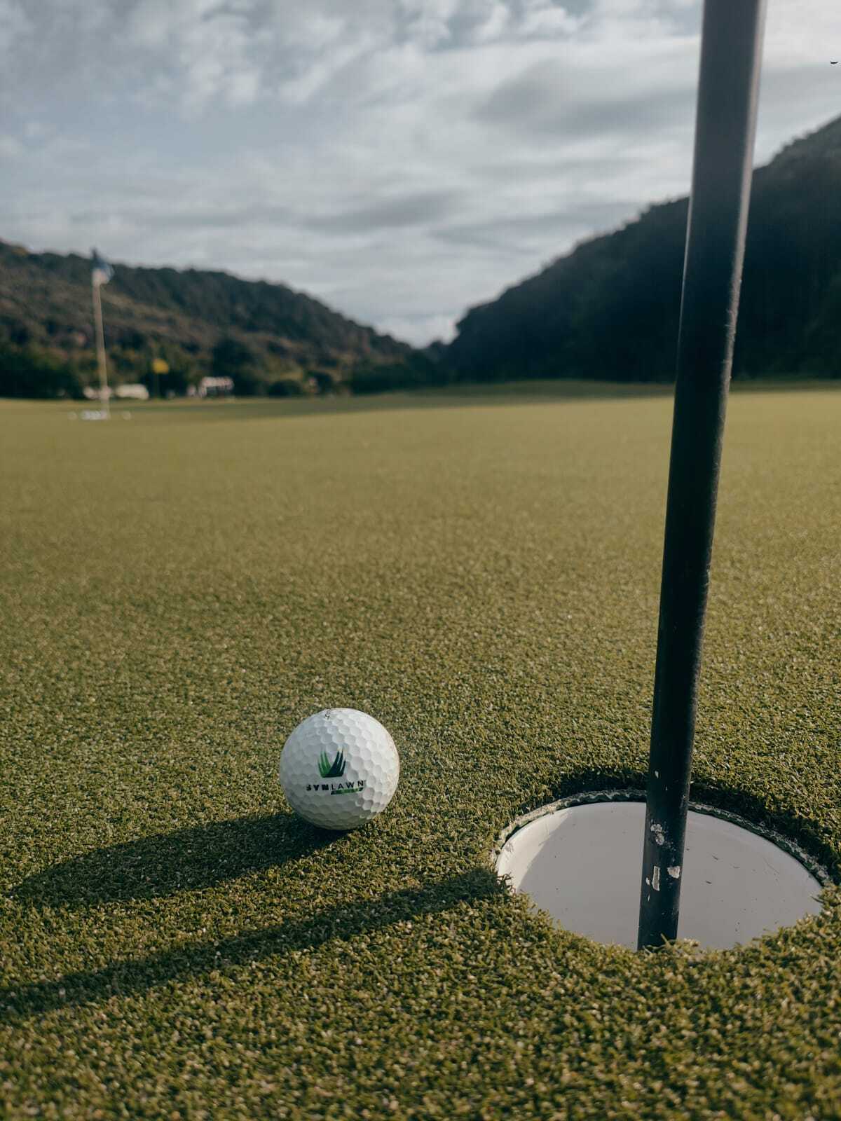 A Synlawn golf ball rests beside the hole on a synthetic putting green with hills and cloudy sky in the background.
