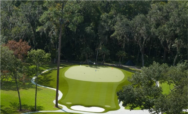 A SYNLawn putting green with smooth synthetic turf, pale bunkers and trees surrounding the curved layout.