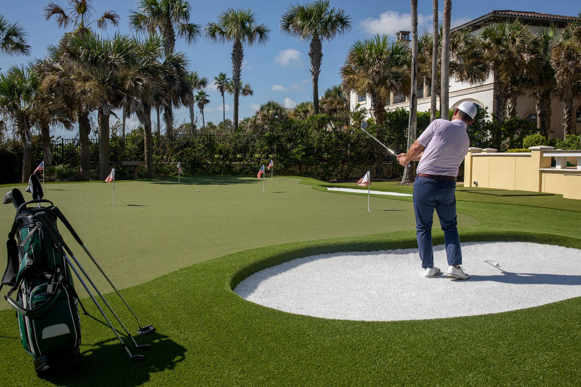 A golfer practises on a custom putting green with synthetic turf, a bunker feature and several flagsticks under palm trees.